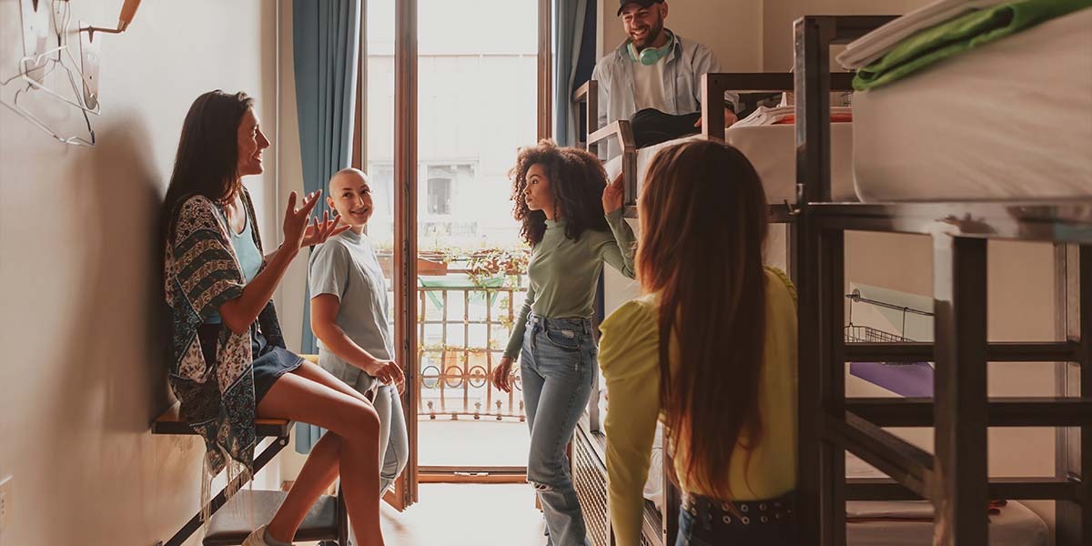 A group of five young people interact joyfully in a sunlit room with bunk beds. One person talks animatedly, while others listen and smile, creating a lively and warm atmosphere.