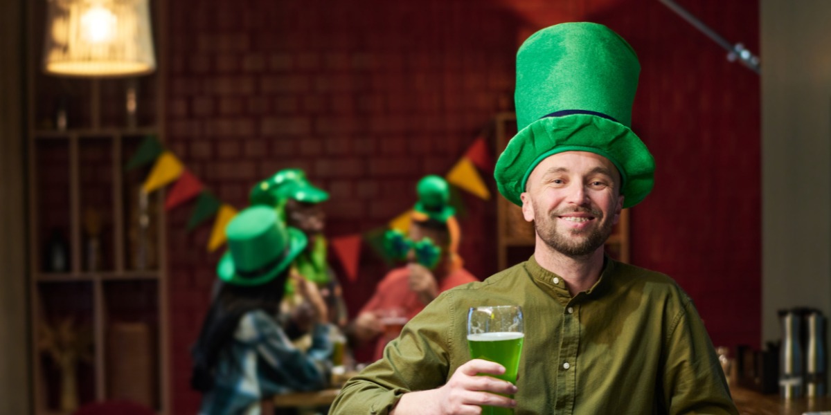 A smiling man in a green leprechaun hat holds a glass of green beer, celebrating St. Patrick's Day. Festive crowd and colorful bunting in the background.