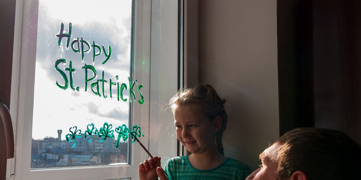 A child in a green shirt smiles while painting "Happy St. Patrick's" with shamrocks on a window. Sunlight streams through, creating a cheerful mood.