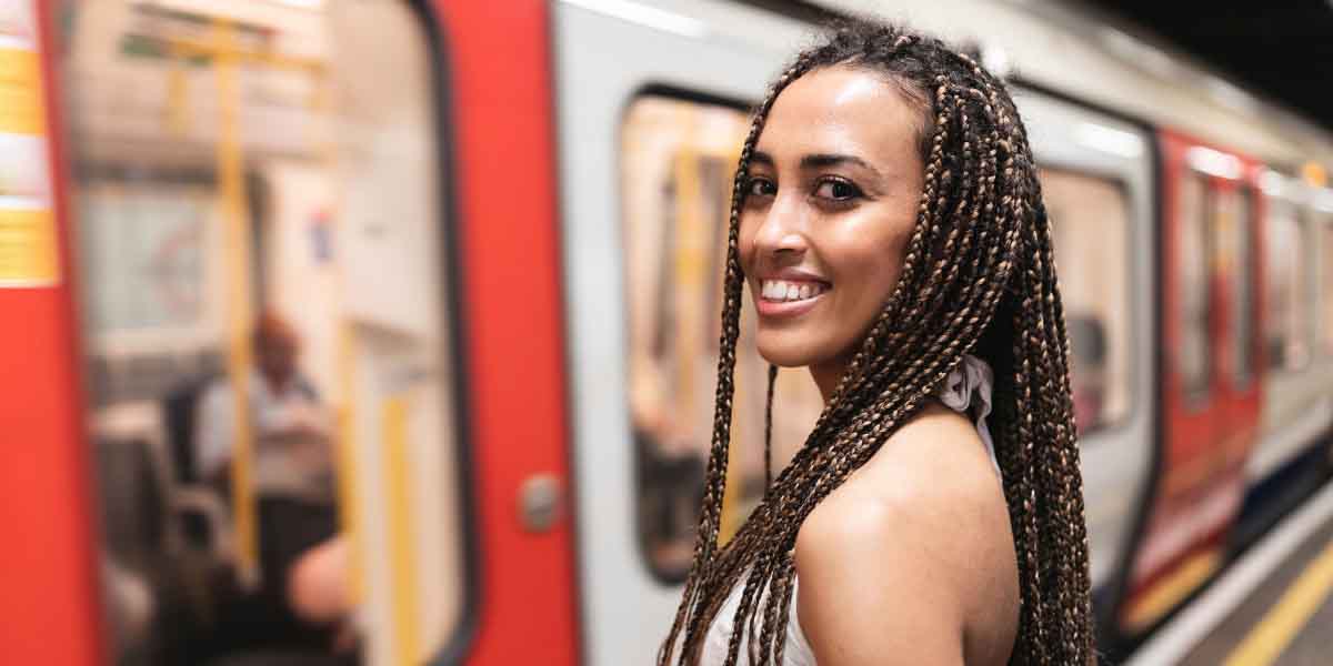 A woman with braided hair smiles at the camera while standing on a train platform. A red train is blurred in motion behind her, conveying energy and travel.
