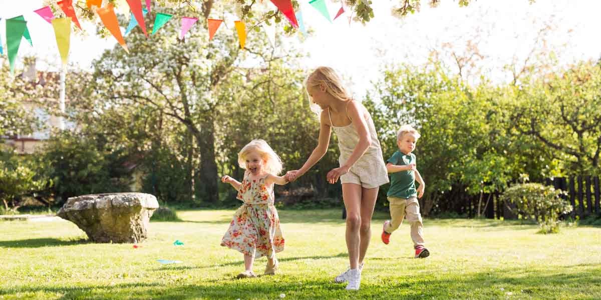 A woman and two children play joyfully in a sunlit garden. Colorful bunting hangs above, creating a festive atmosphere. Bright and cheerful scene.