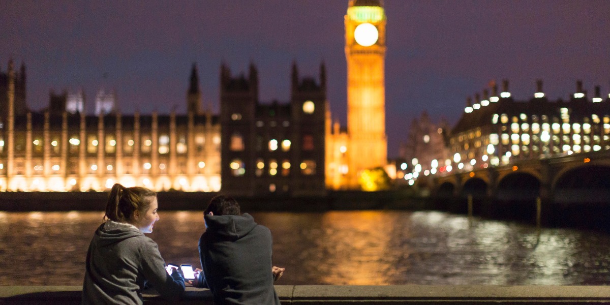 A young couple leans on a riverside wall at night, gazing at the illuminated Big Ben and Houses of Parliament, reflecting on the Thames. A serene, romantic atmosphere.