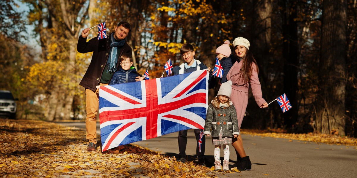 A family of five stands on a leaf-covered road, holding a large Union Jack and small flags. It's a bright autumn day, conveying joy and togetherness.