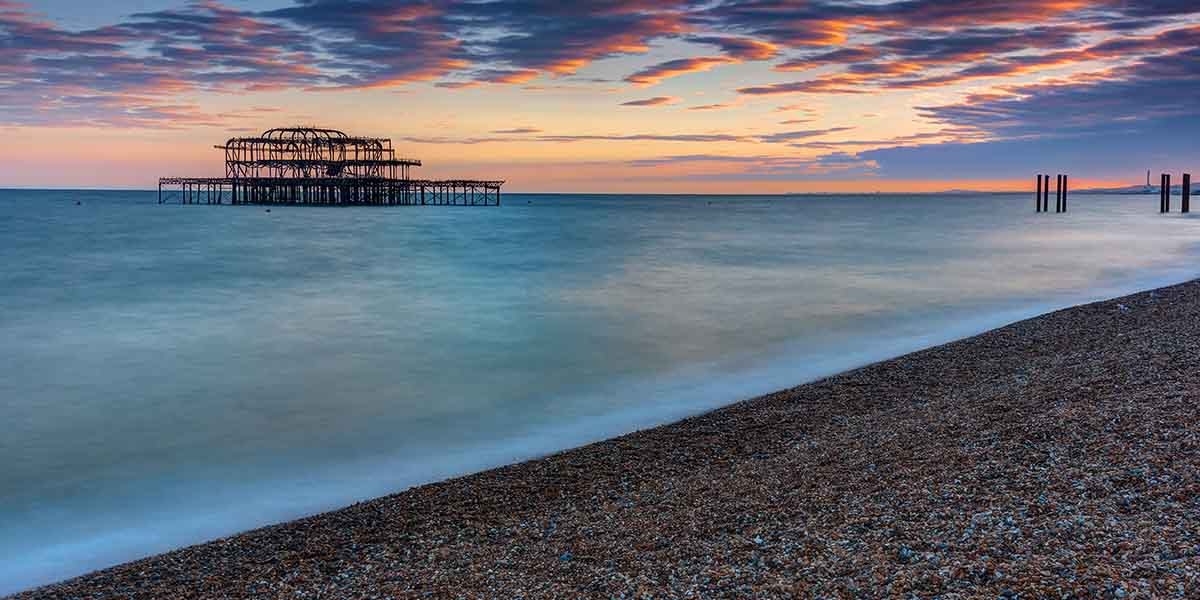 Sunset over calm sea with a pebbled beach in the foreground. An old pier structure stands in the water. The sky is vibrant with pink and orange hues.