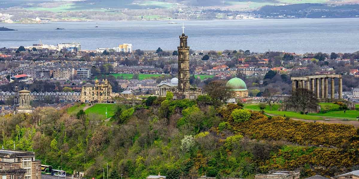 A scenic view of Calton Hill in Edinburgh, featuring lush greenery, historic monuments, and a cityscape backdrop with a serene water body beyond.