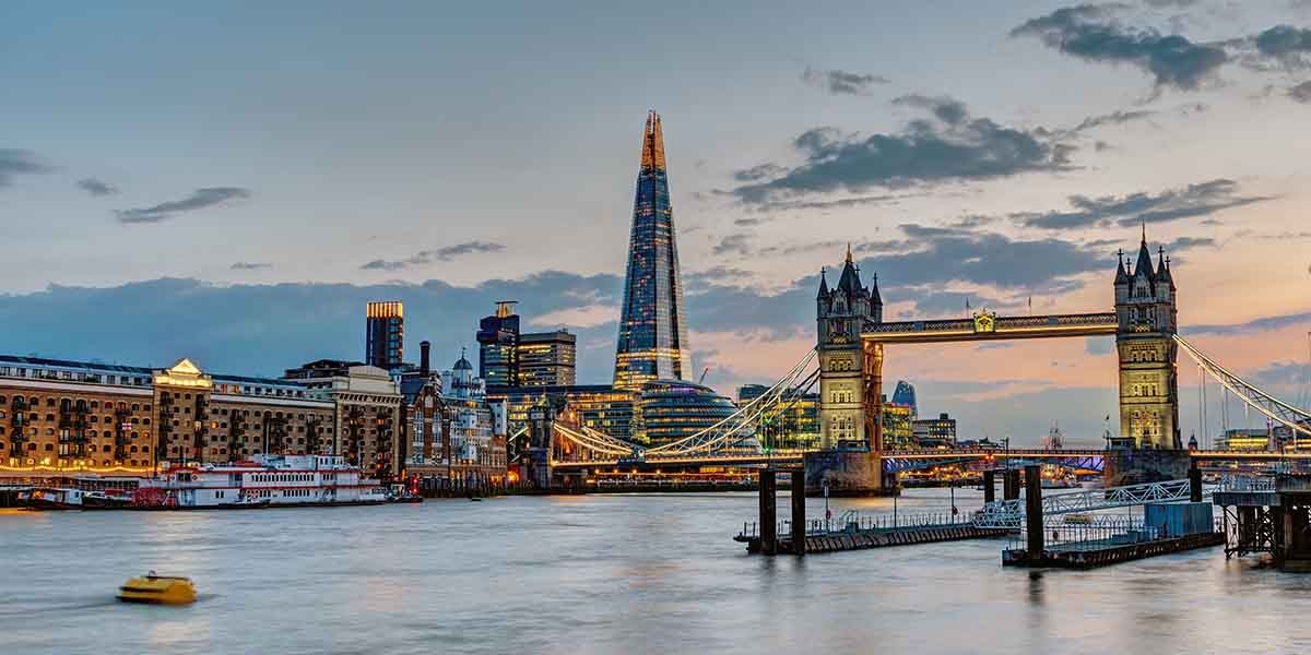 London's skyline at sunset with the Shard and Tower Bridge. The River Thames reflects vibrant colors, creating a serene and iconic urban scene.