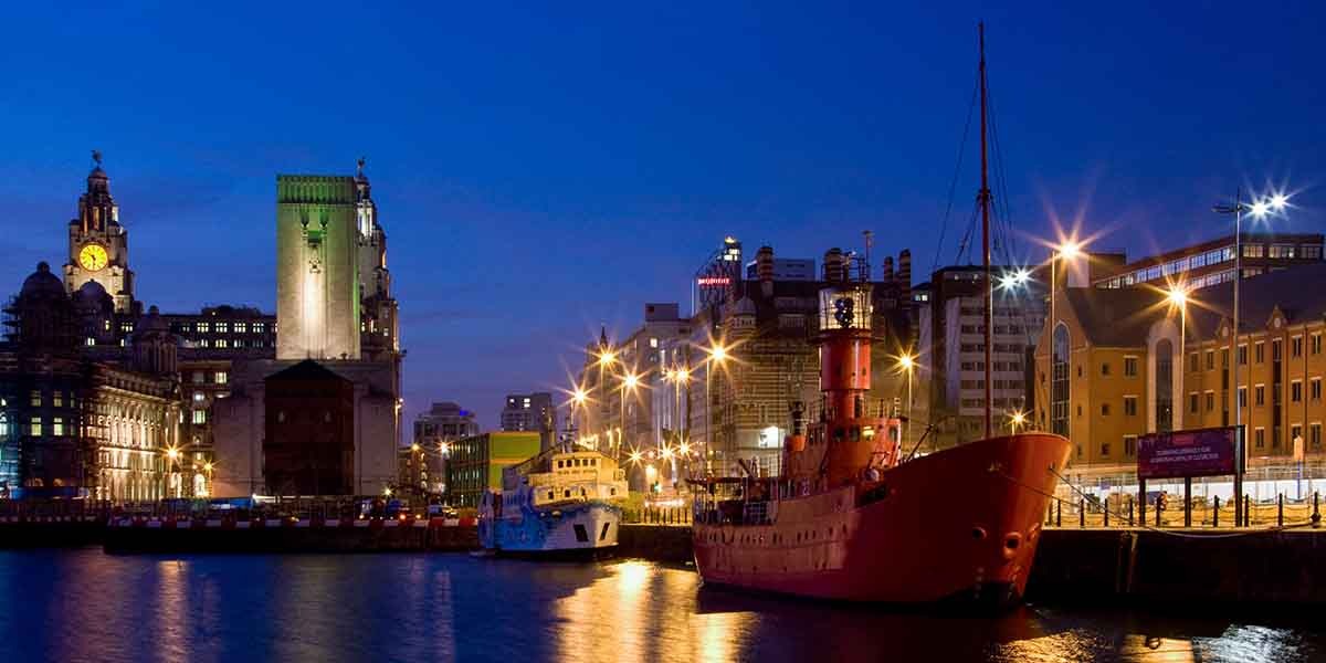 Nighttime view of Liverpool's waterfront. A red ship is docked near lit buildings, including the iconic Royal Liver Building, under a deep blue sky.