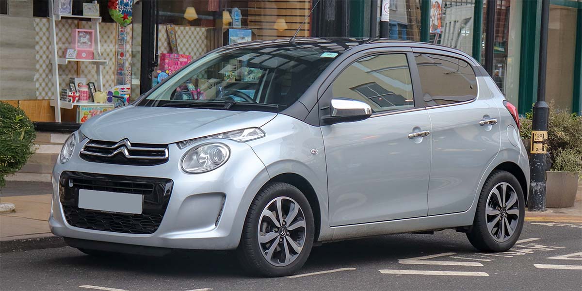 A silver Citroën compact car is parked on a street near a shop with colorful displays in the window. The car's modern design is sleek and polished.