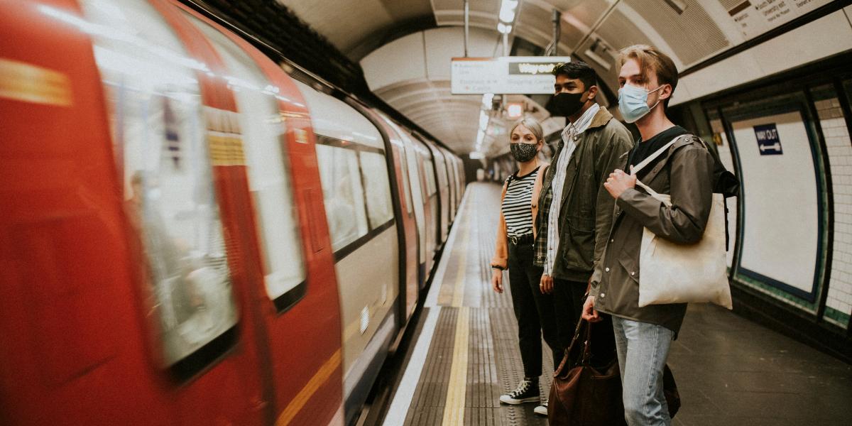 Three people wearing masks wait on a subway platform as a train approaches. The scene feels tense and cautious amid a dimly lit underground setting.