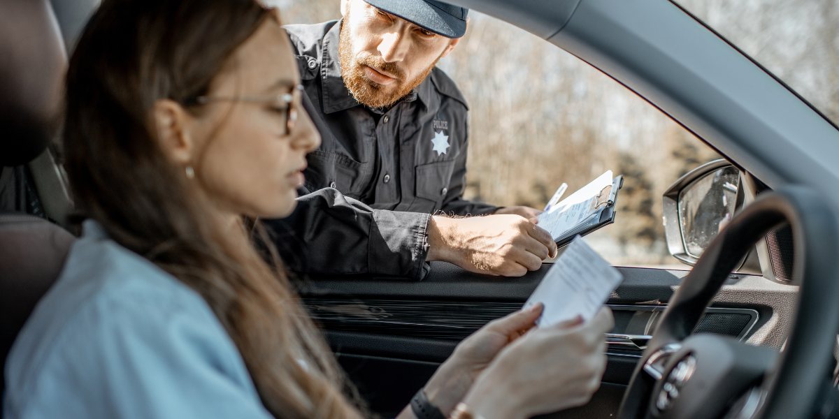 A police officer leans at a car window, examining a driver's documents with focus. The driver, seated and holding papers, appears attentive.