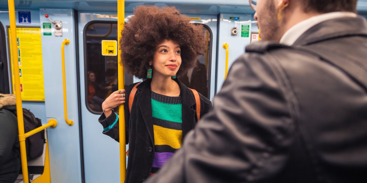A woman with curly hair, wearing a colorful striped sweater, smiles while holding a subway pole. A man in a leather jacket faces her, setting a cheerful subway scene.