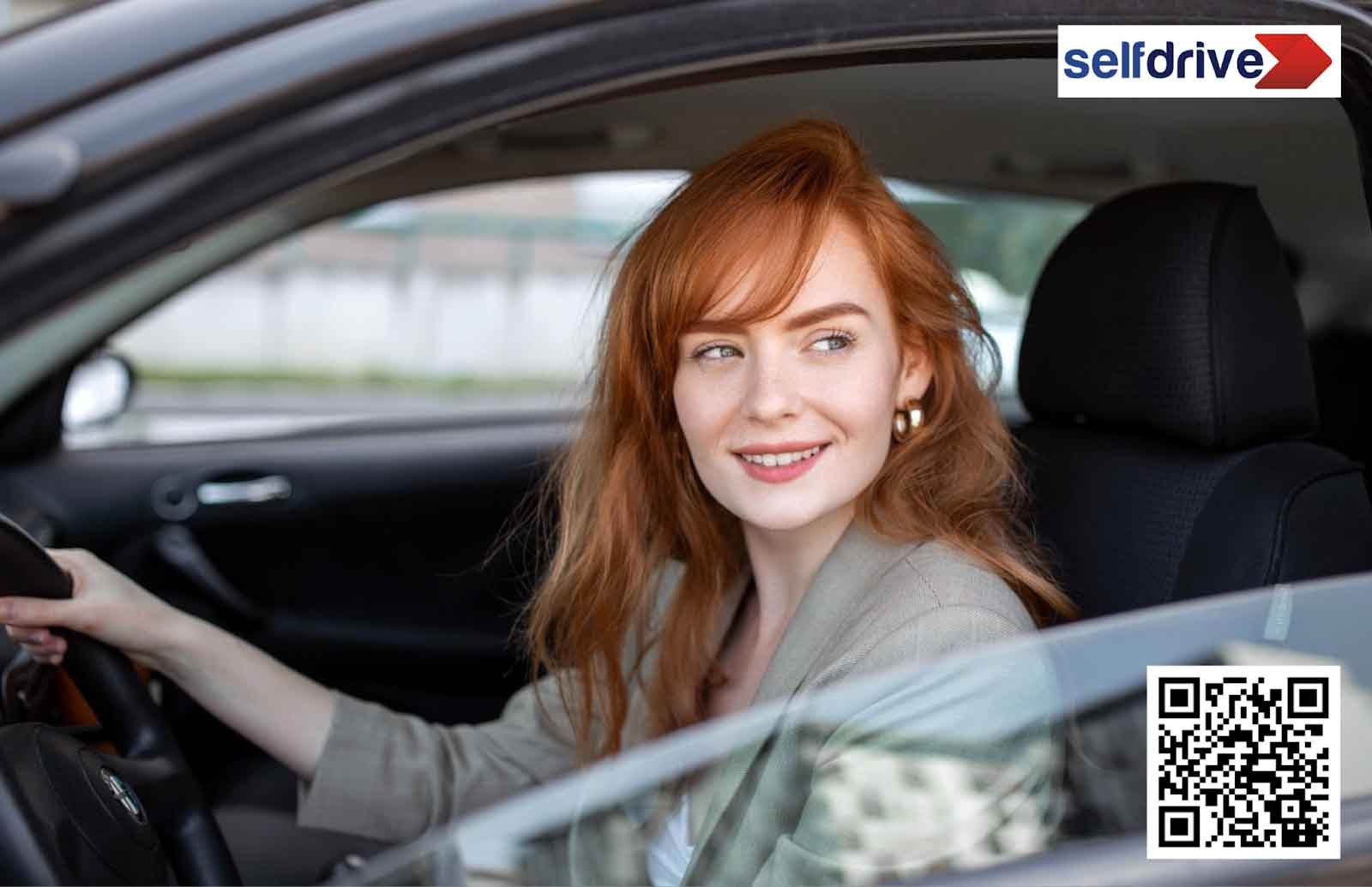 A smiling woman with red hair is sitting in a car, looking out the window. The car advertises 